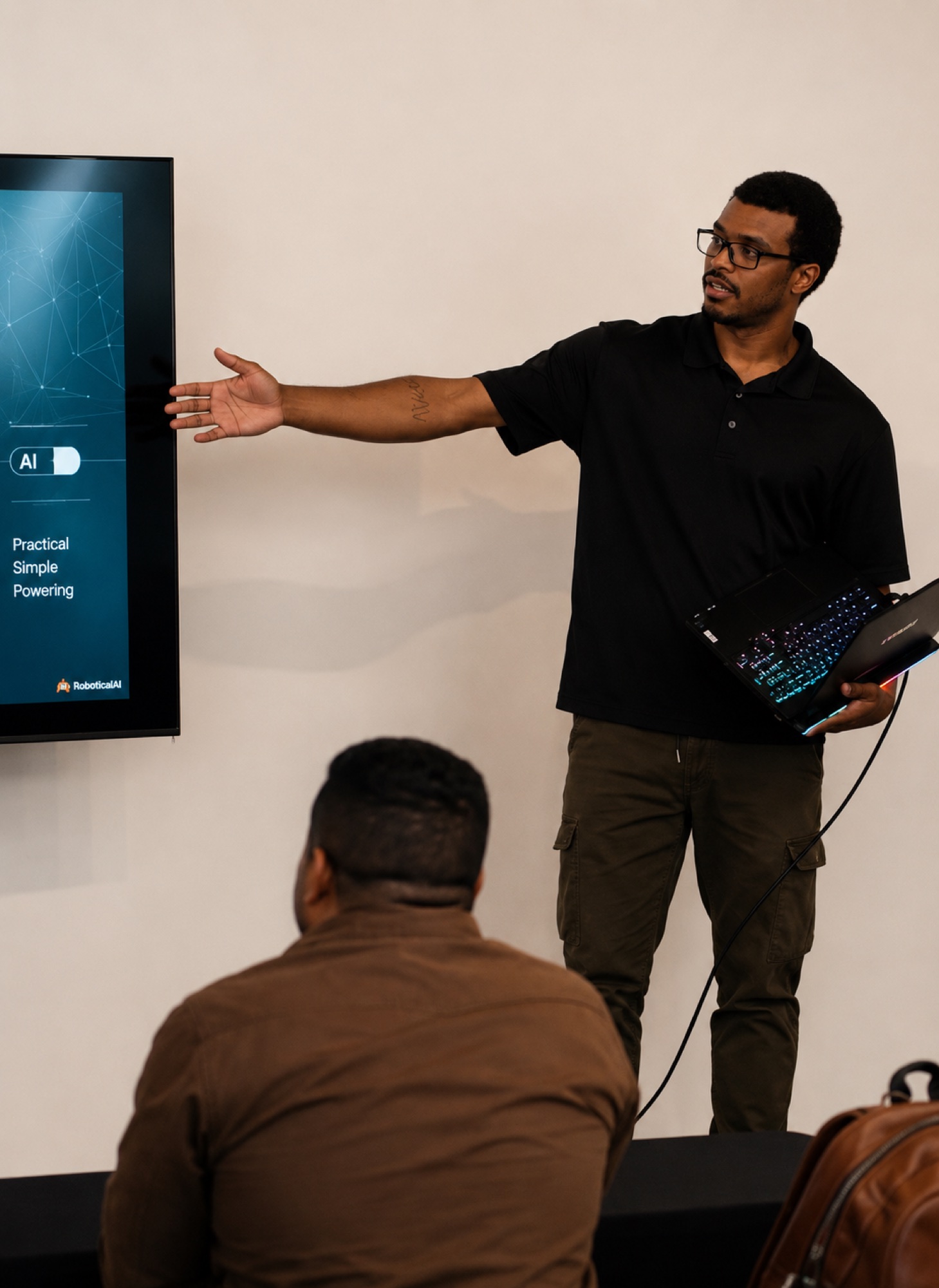A presenter holding a laptop and pointing to a screen while demonstrating an AI tool to the room